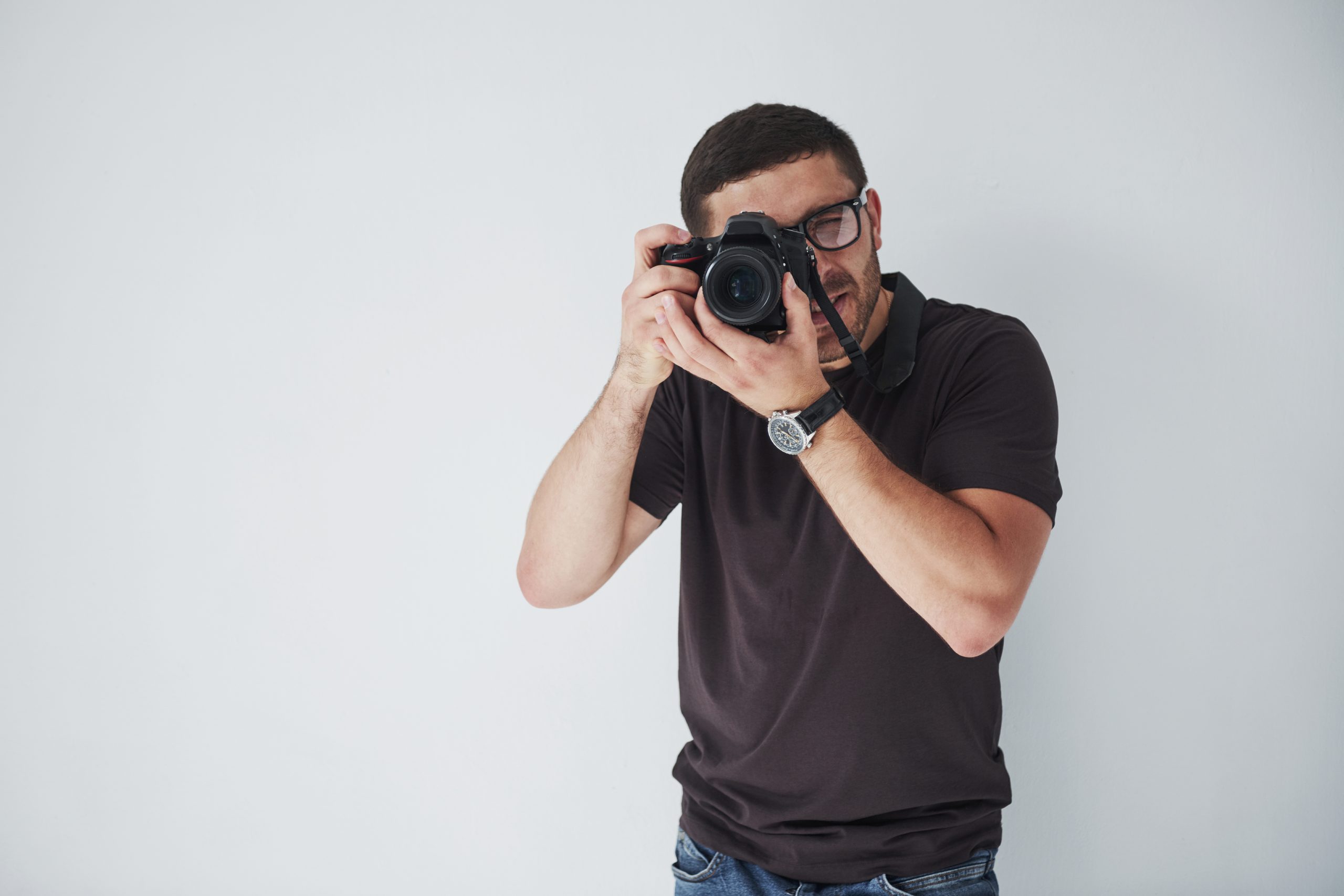 A young hipster man in eyepieces holds a DSLR camera in hands standing against a white wall background.